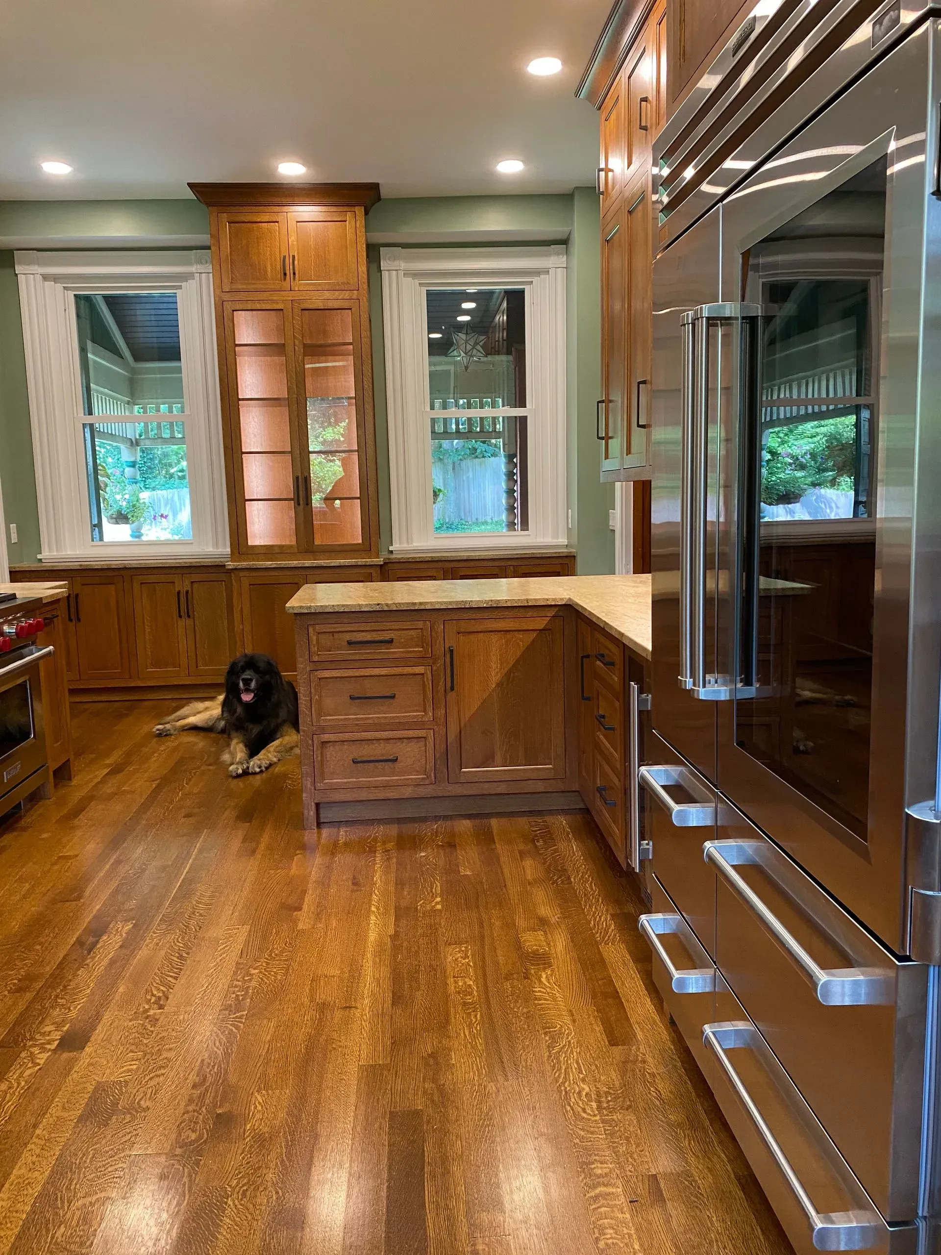 A kitchen with wood cabinets and floors, large stainless steel refrigerator, windows, and a dog.