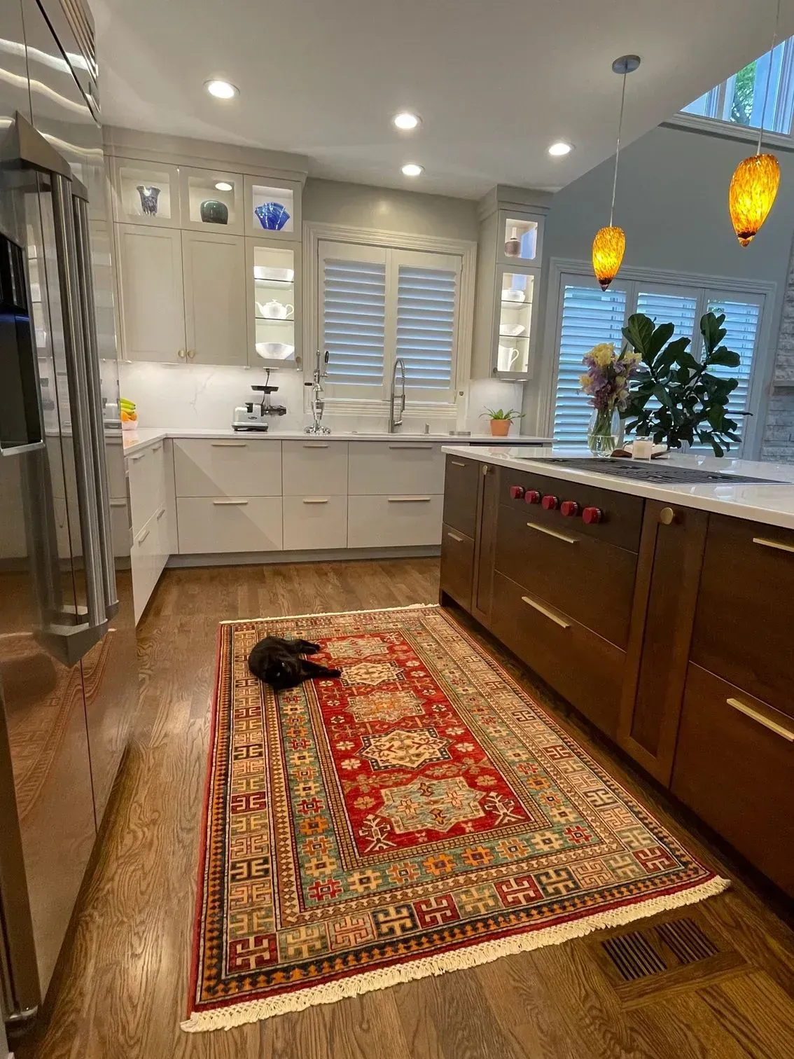 A kitchen with a rug and a black cat. The space has wood cabinets, white countertops, and warm lighting.