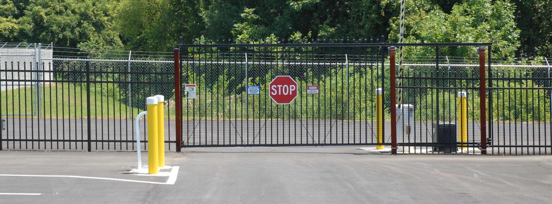 Black metal gate with a red stop sign and yellow bollard at a fenced entrance.