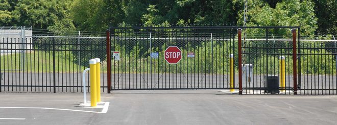 Black metal gate with a red stop sign and yellow bollard at a fenced entrance.