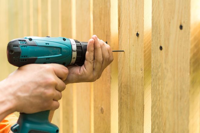 Hands using a teal drill to drive a screw into a wooden fence panel