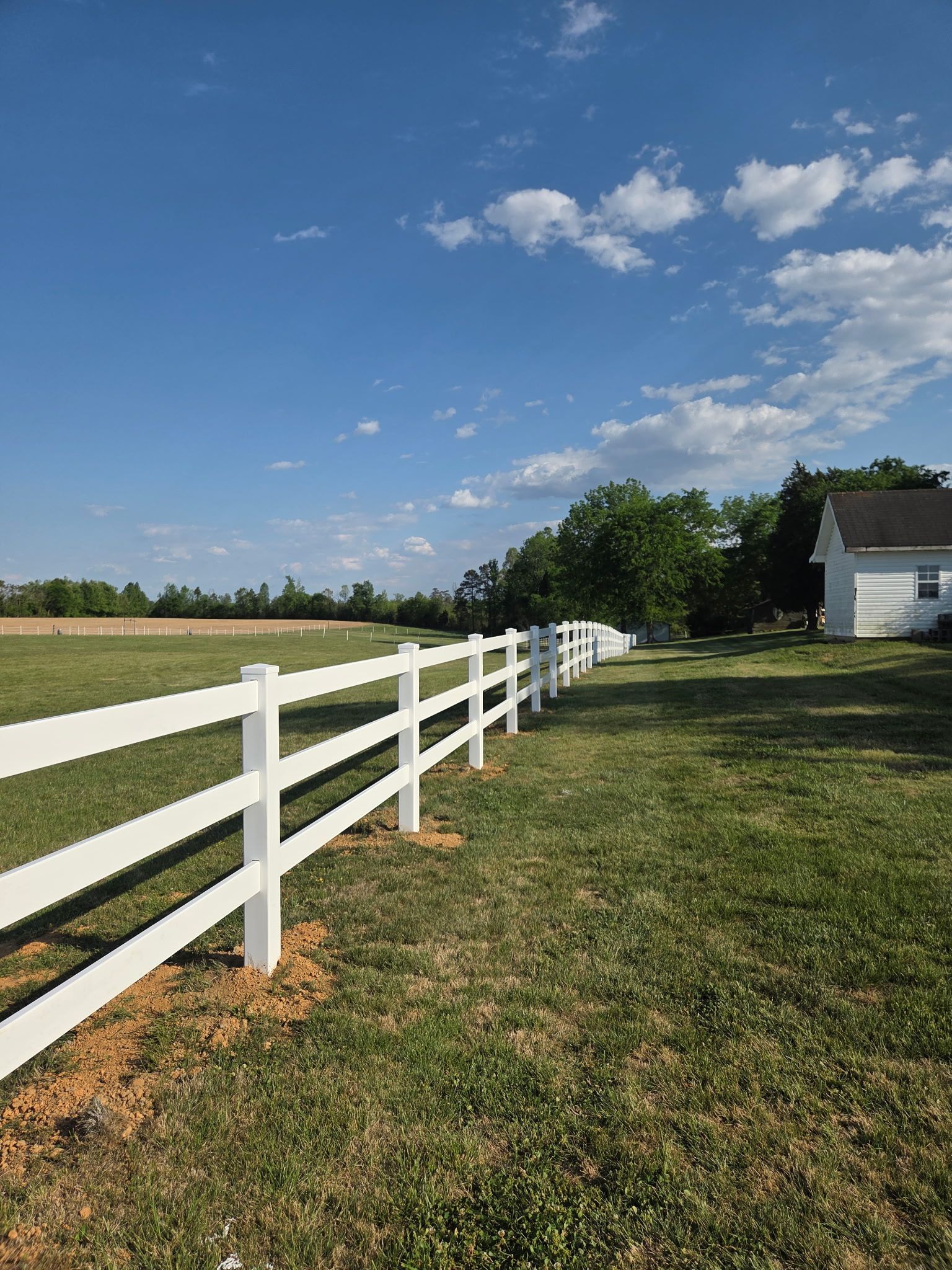 White fence along a grassy field under a blue sky, with trees and a small house in the distance
