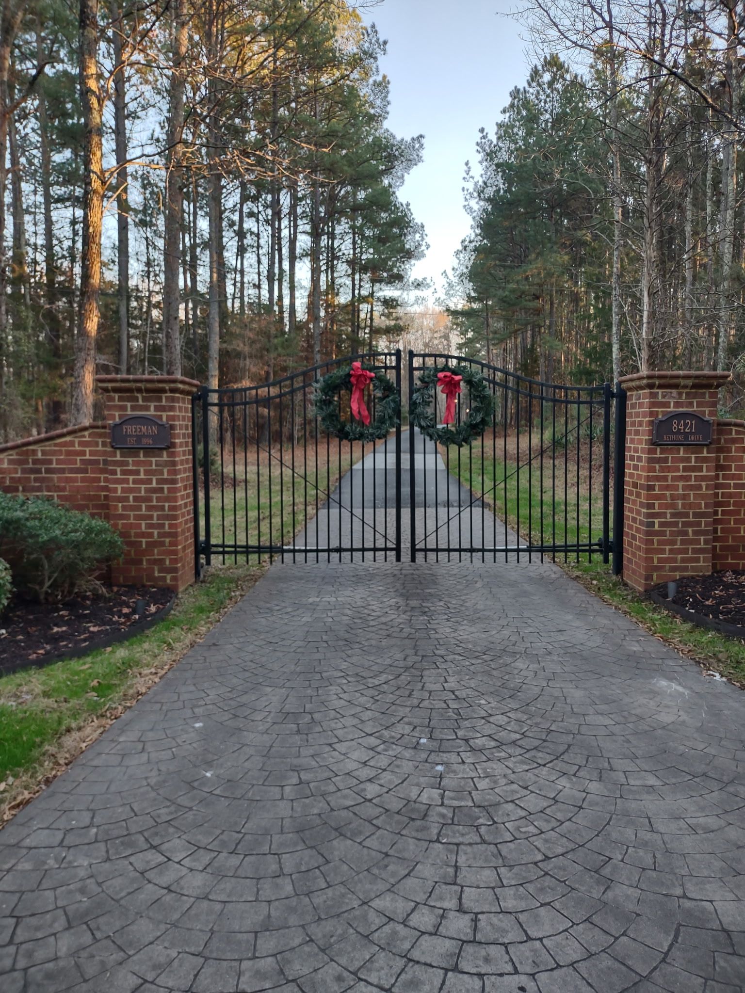 Gated driveway with holiday wreaths, brick pillars, and tall trees lining the entrance.