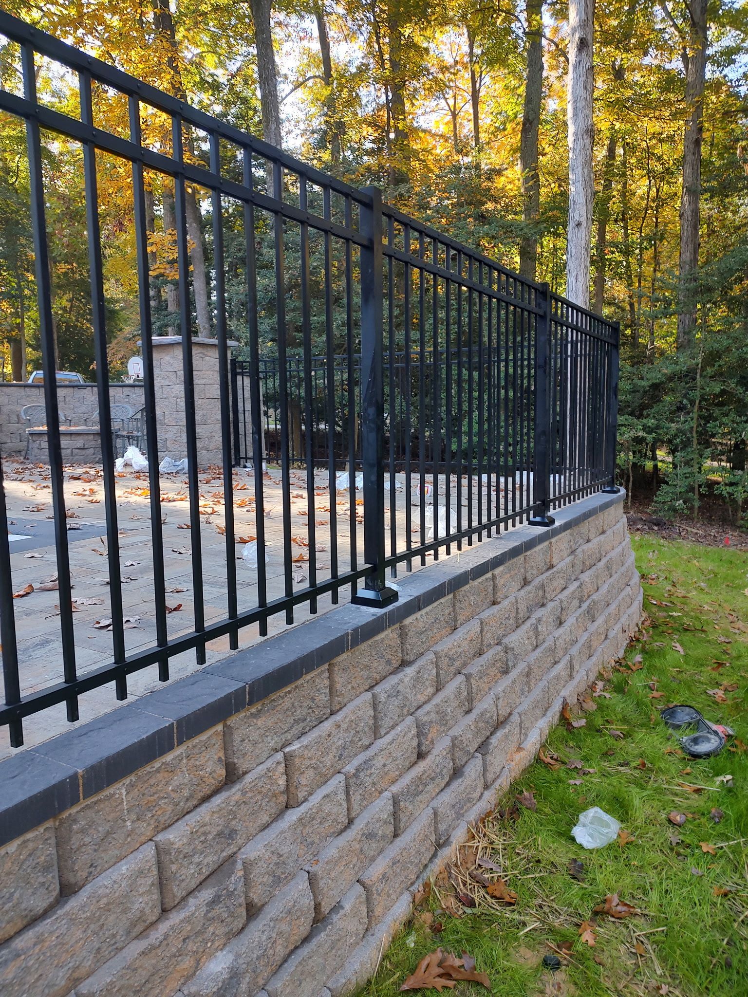 Black metal fence atop a stone retaining wall beside a grassy yard and wooded area.