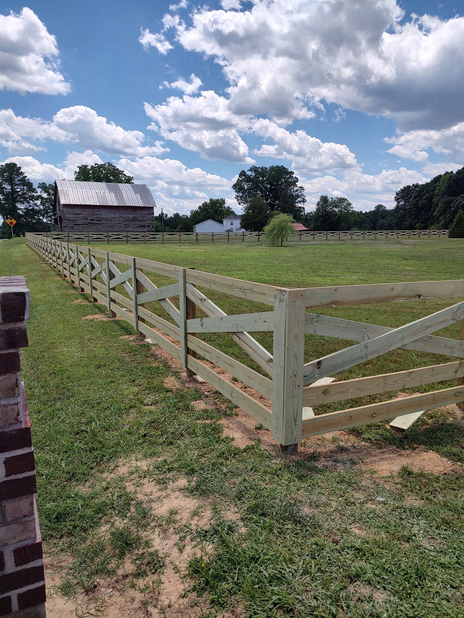 White wooden fence around a grassy field under a cloudy blue sky with a barn in the distance