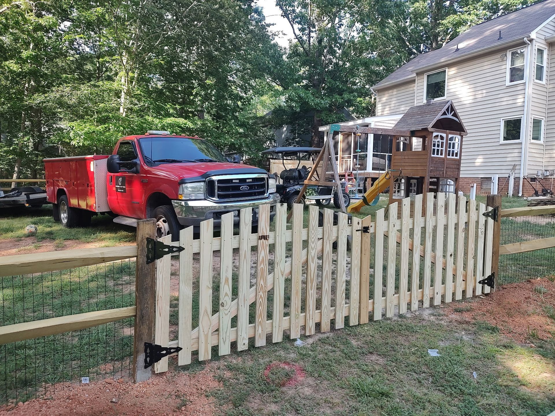 Red pickup truck and trailer beside a white house, viewed through a wooden fence in a yard.