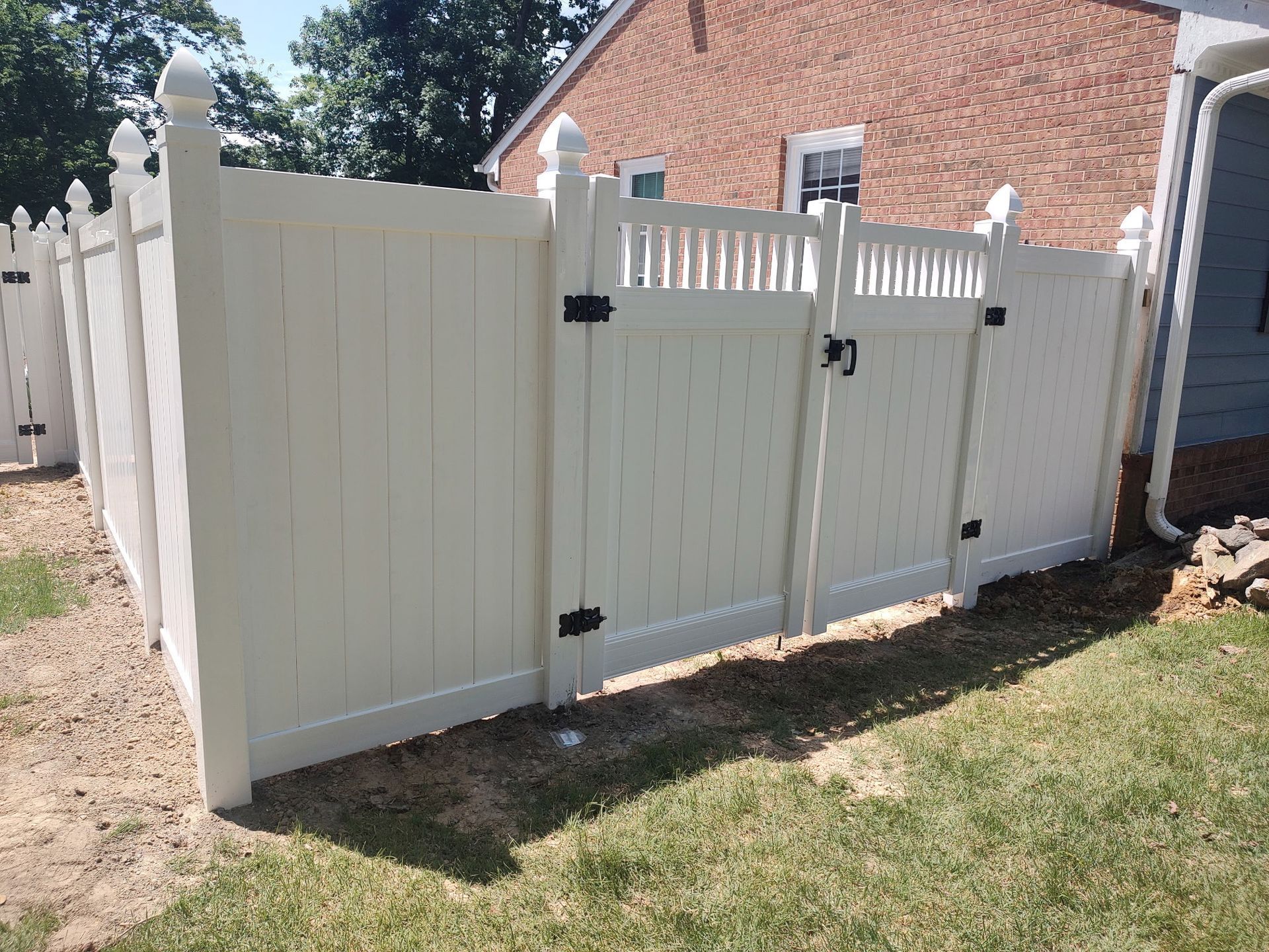 White vinyl fence with a double gate beside a brick house on a sunny yard