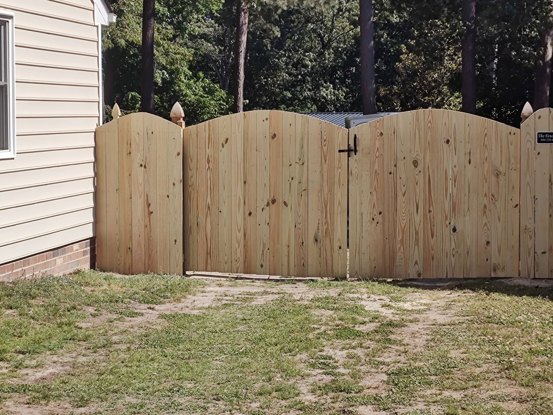 Wooden backyard gate in a fenced yard beside a beige house, with grass and trees behind it.