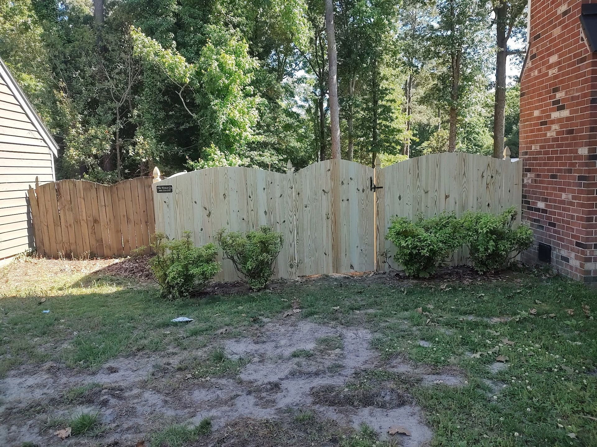 Backyard with wooden fence, shrubs, patchy grass, and a brick wall on the right.