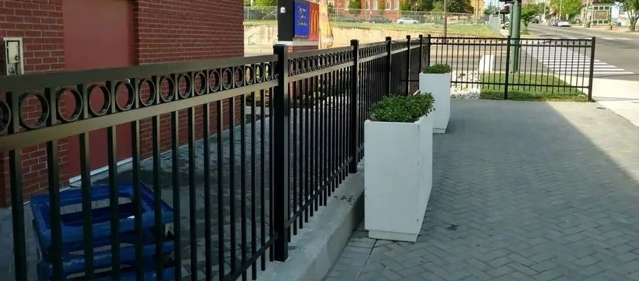Black metal fence beside a sidewalk with a white planter and brick building entrance