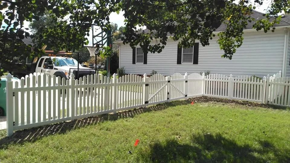 White picket fence around a green yard beside a white house, with a tree casting shade.