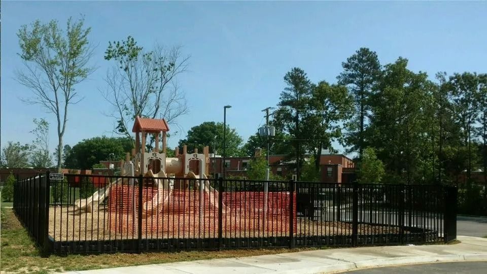 Fenced playground with red play structure and trees under a clear blue sky