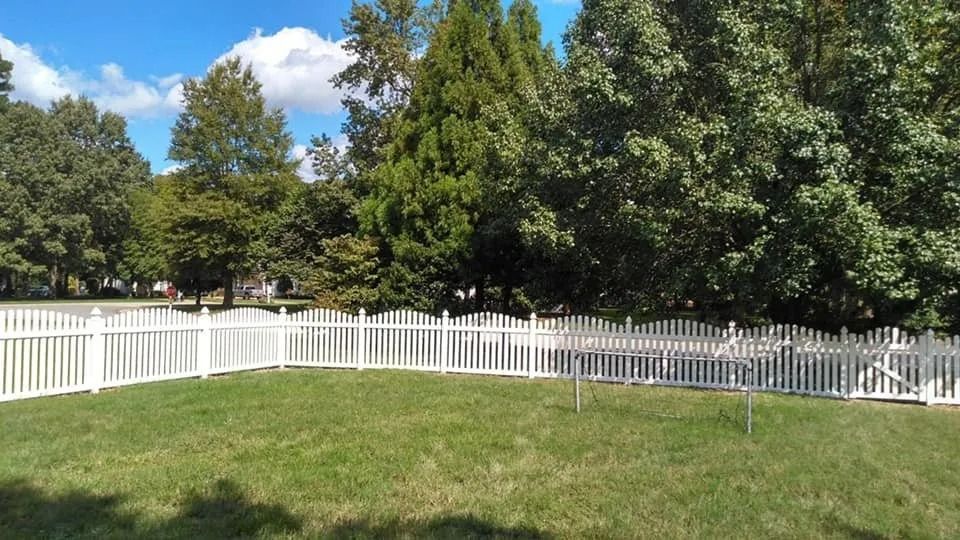 White picket fence curving around a green lawn with tall trees under a blue sky with clouds
