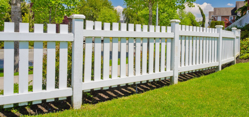 White picket fence lining a green lawn with leafy trees in the background