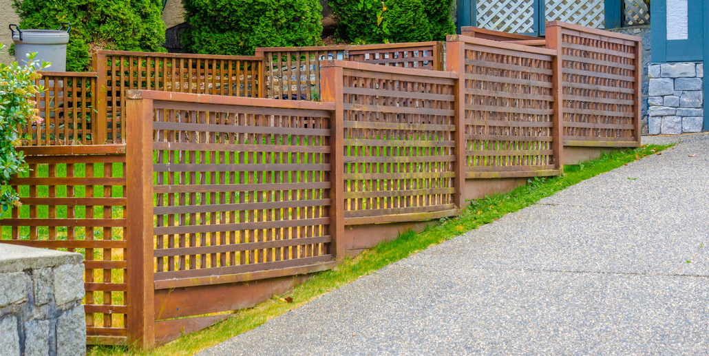 Wooden lattice fence along a gravel driveway beside a green garden and stone wall