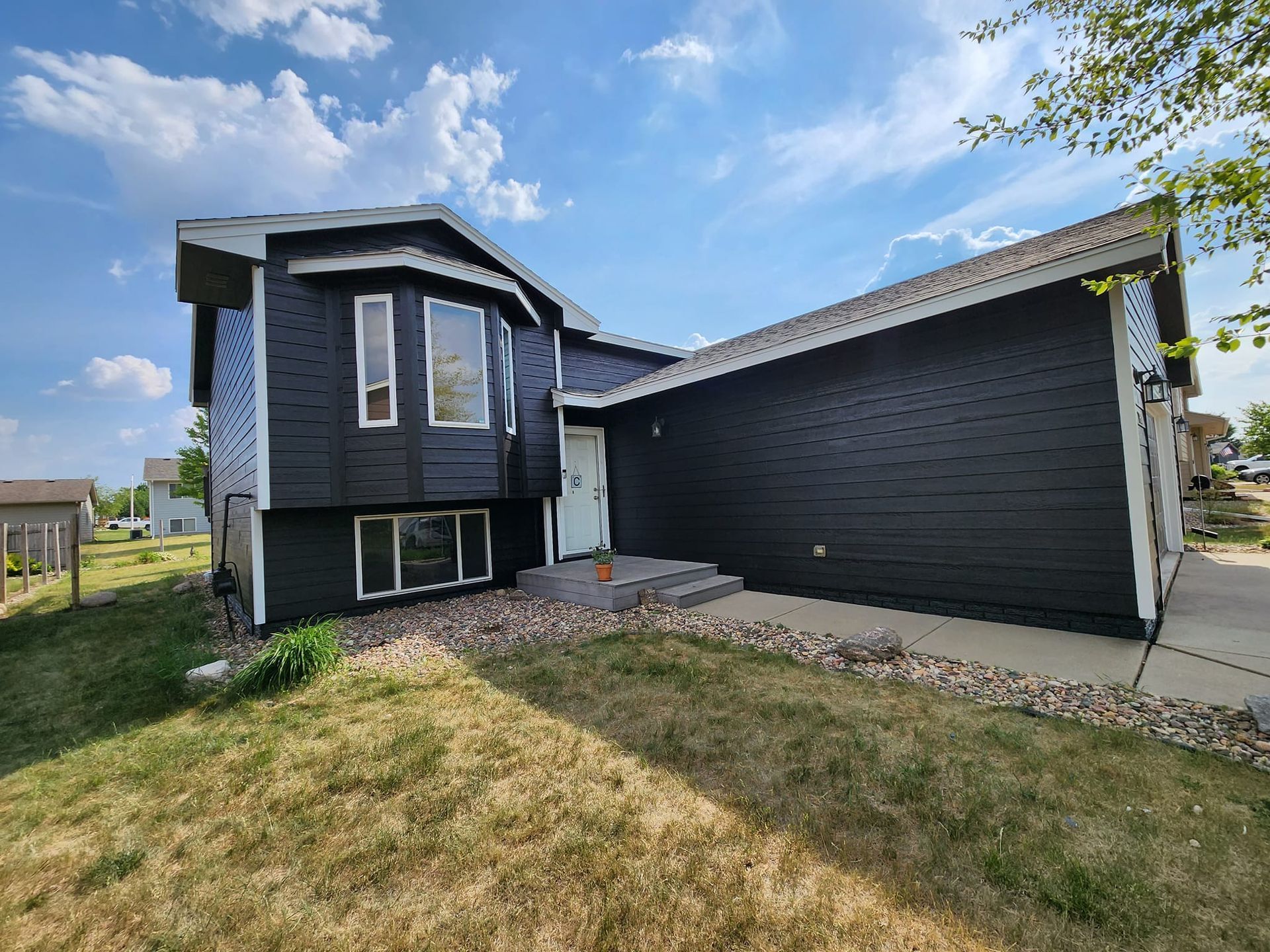 Black house with white trim, bay window, front door, and dry grass on a sunny day.