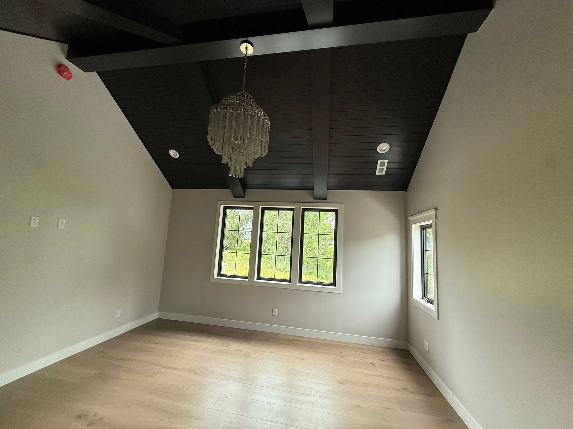 Empty room with dark ceiling beams, chandelier, windows, and light-colored walls and floor.