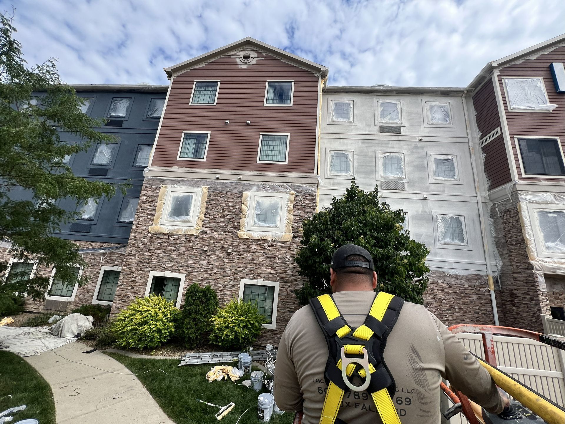 Construction worker with safety harness, looking at a building exterior under renovation with brown and stone textures.