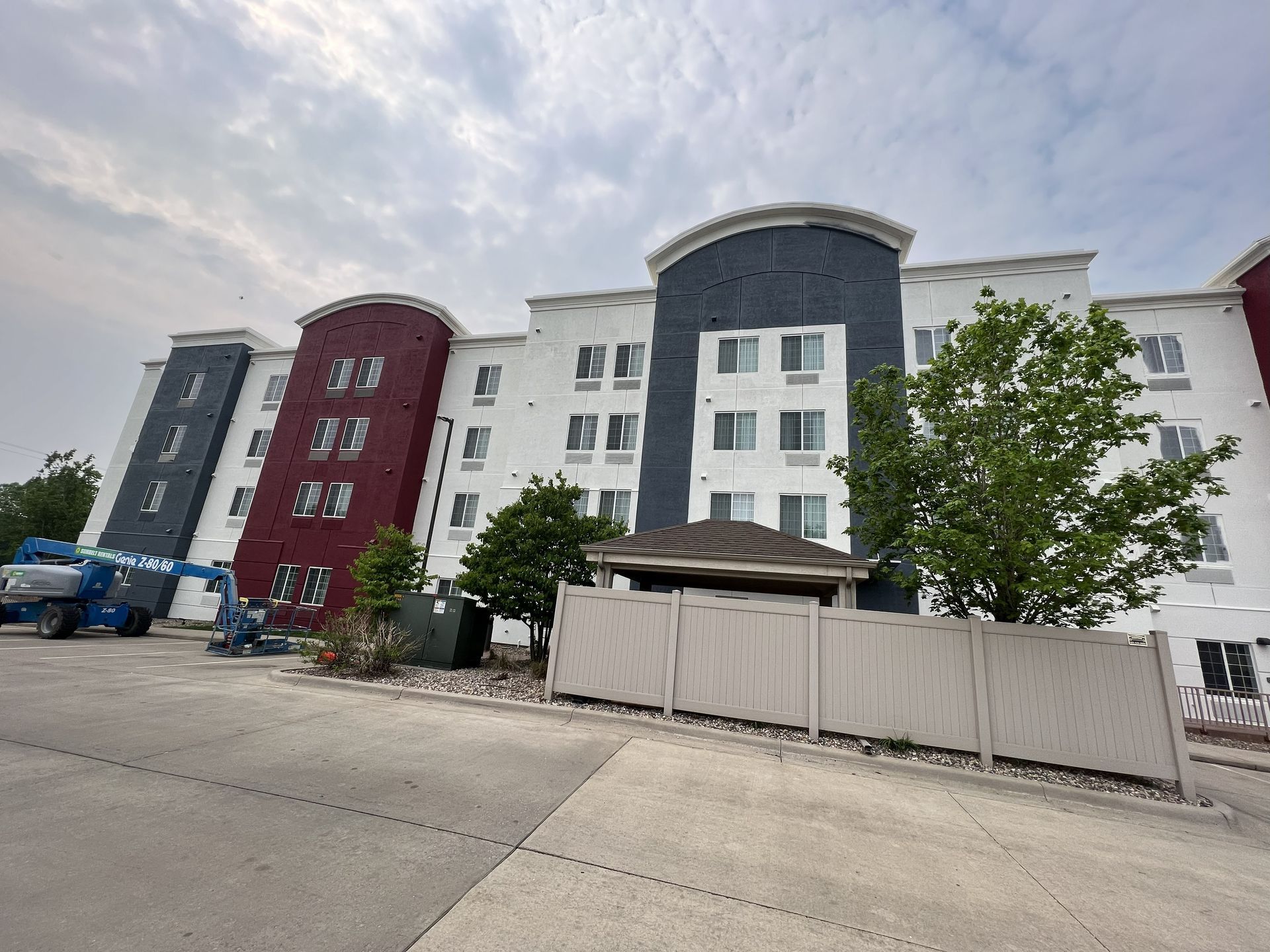 Multi-story building with white, red, and blue facade, cloudy sky. Parking lot and small shed in front.