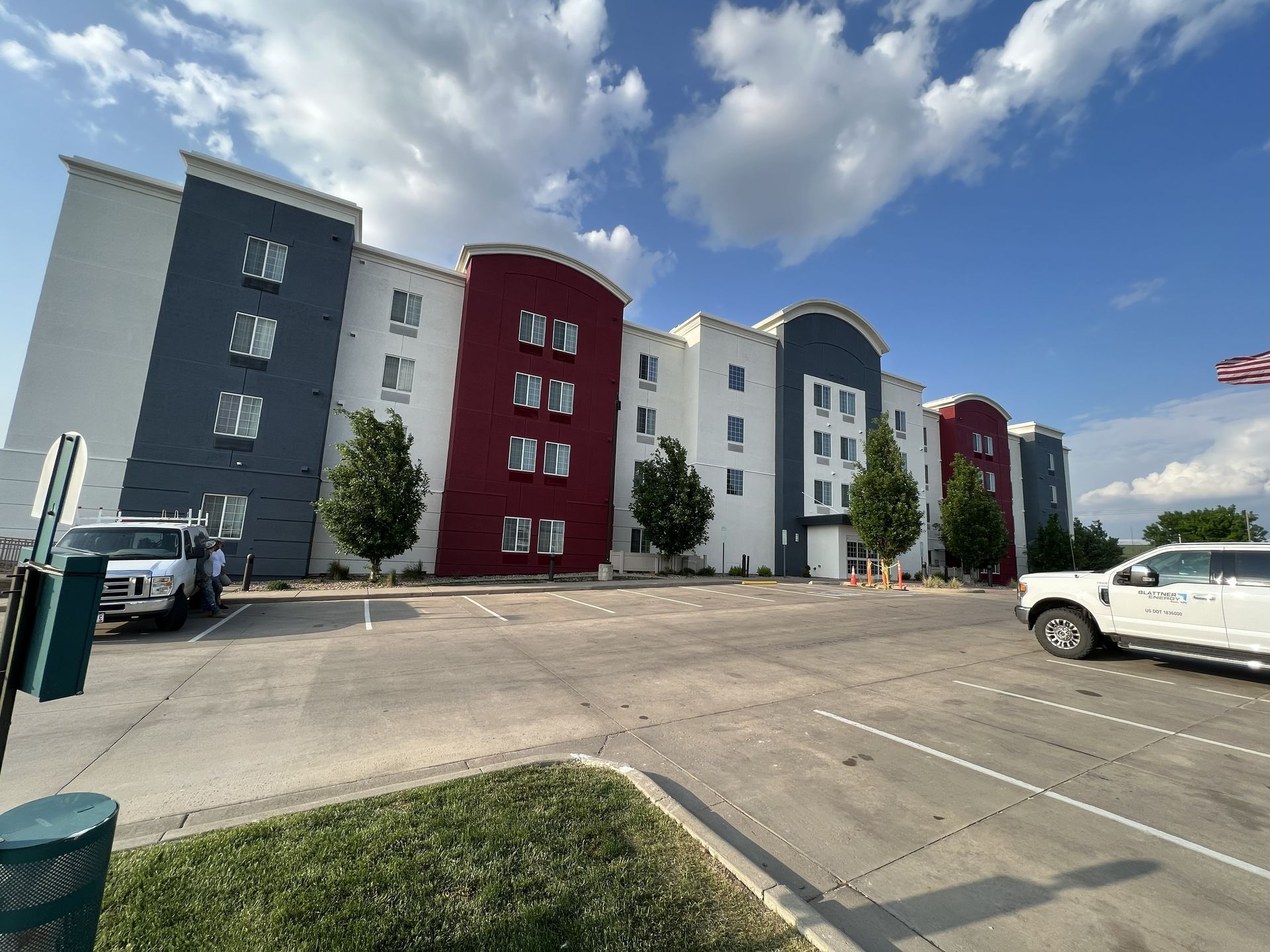 A three-story hotel with white, red, and blue exterior. Parking lot with vehicles. Blue sky and clouds.
