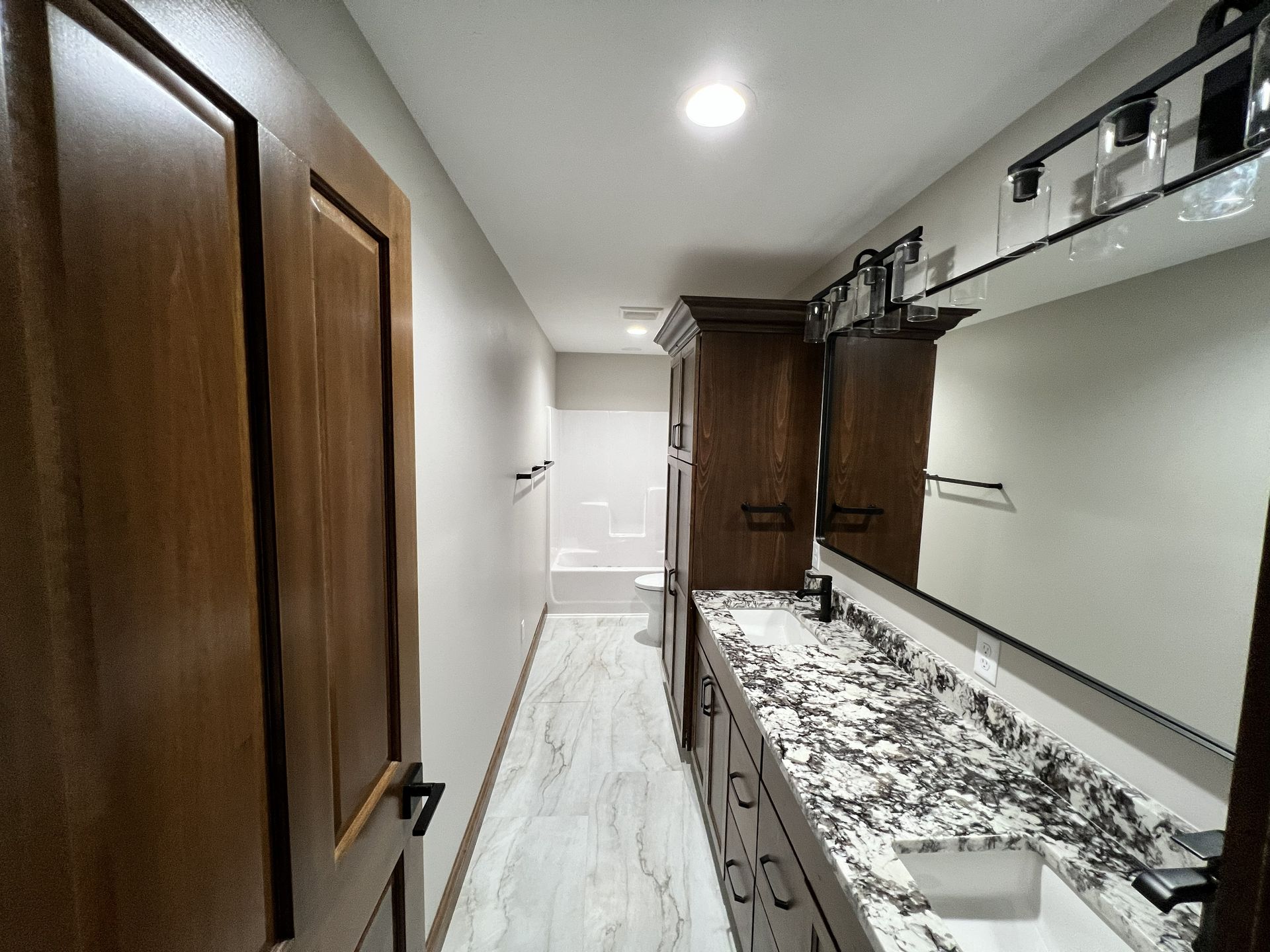 Bathroom with wood door, vanity, and marble countertop. Long and narrow space with light flooring and recessed lighting.