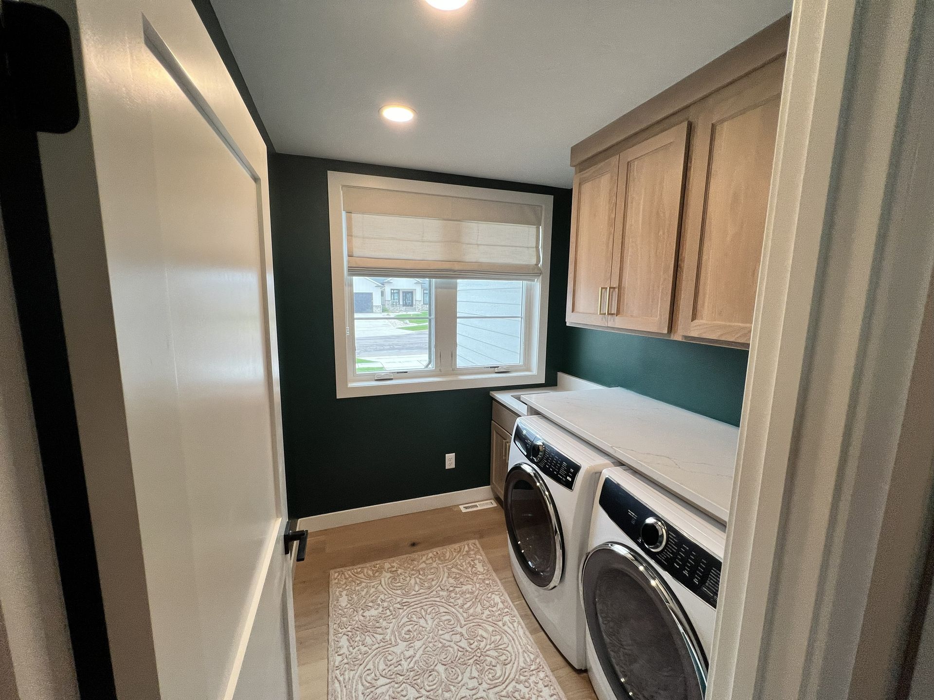 Laundry room with washer, dryer, cabinets, and window. Dark green walls, light wood cabinets, and beige rug.