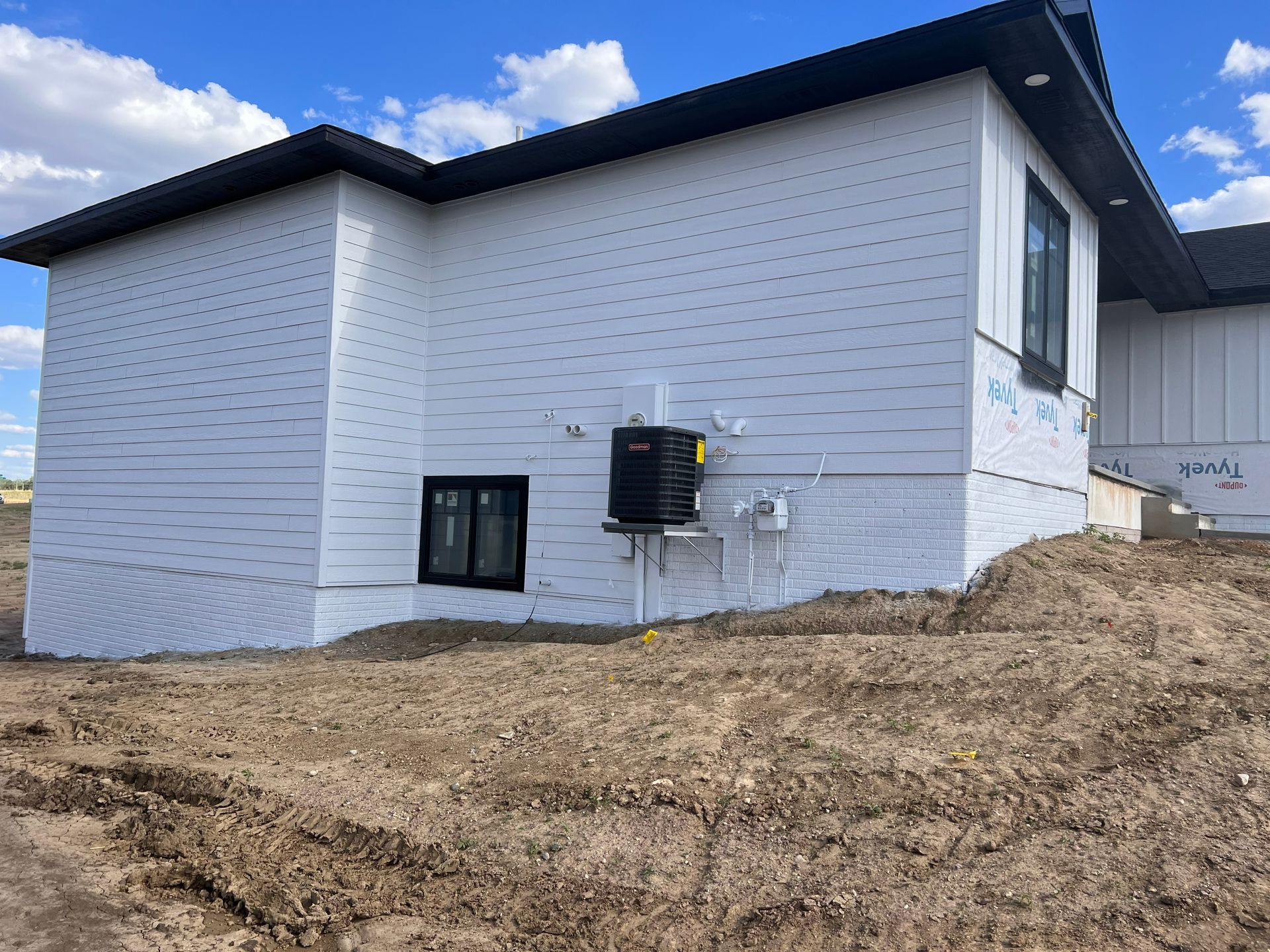 White house under construction with black roof and water tank on the wall, set on a dirt lot.
