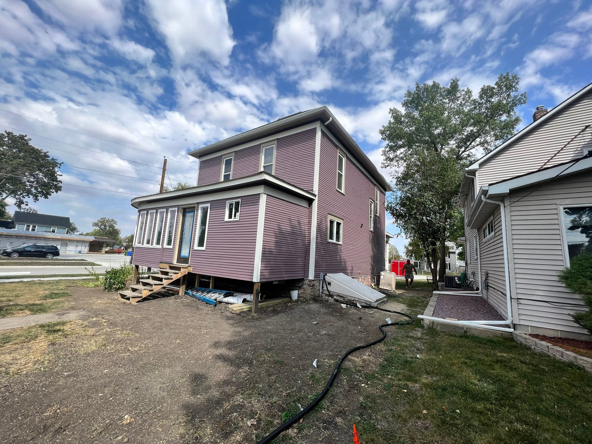 Two-story purple house with screened porch, wooden steps, and adjacent white house under a blue sky.
