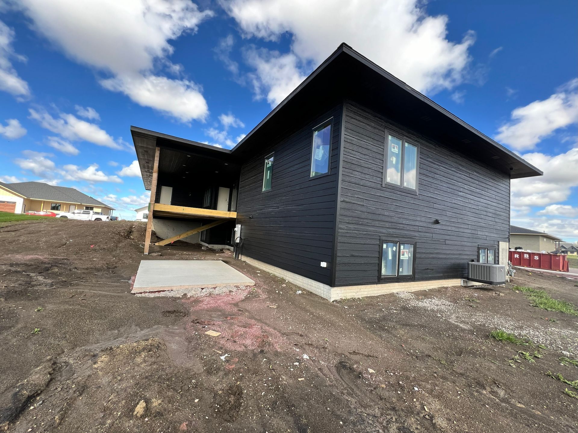 Black-sided house under construction with a deck, windows, and concrete patio, set against a cloudy blue sky.