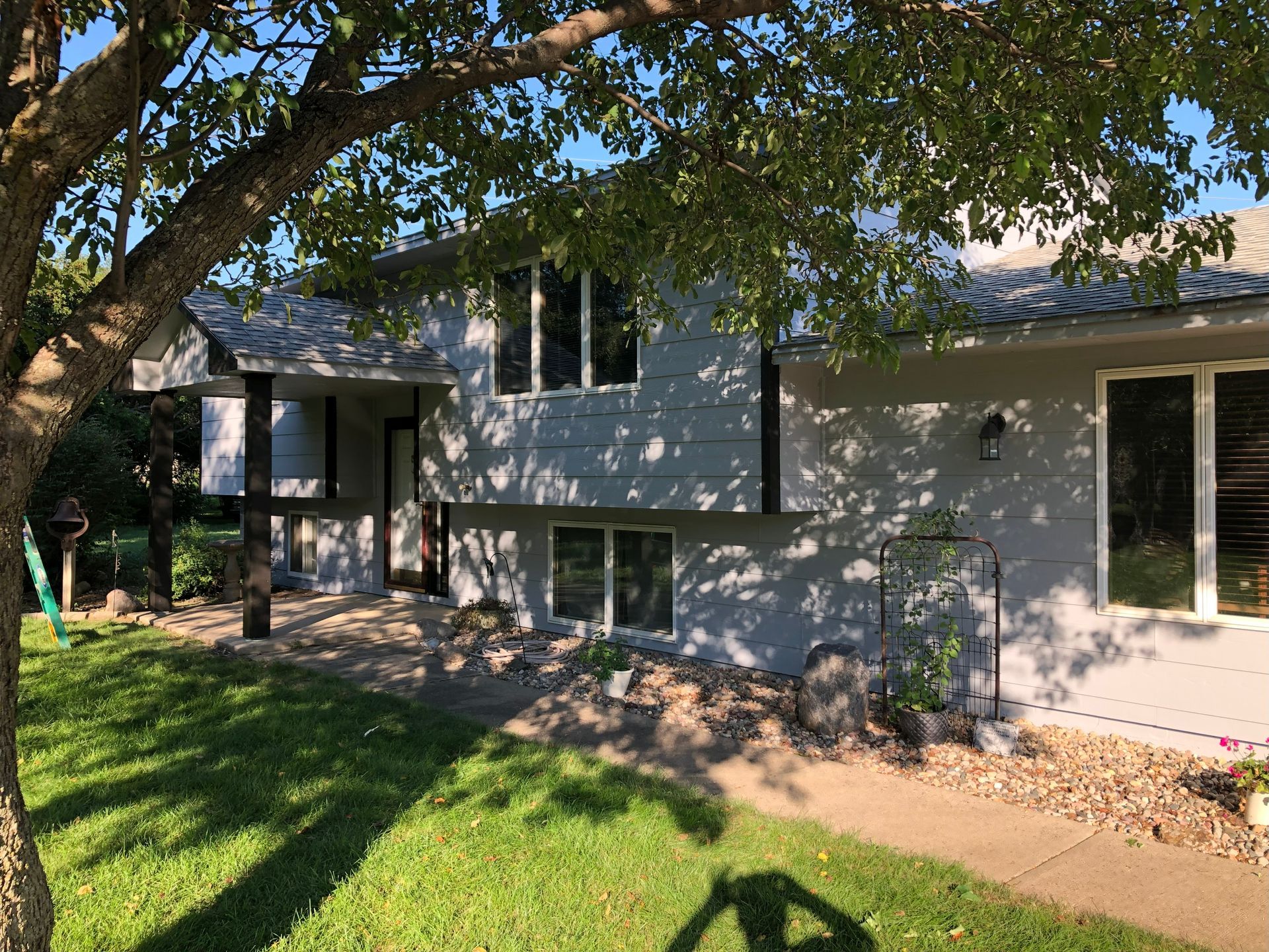 Gray house with black trim, windows, and a tree in the foreground. Sunny day, green grass, and sidewalk.