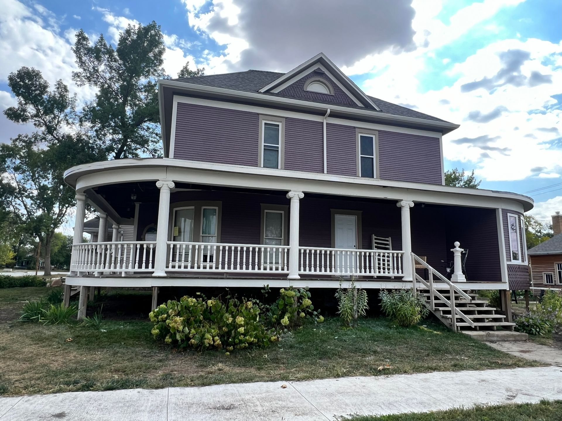 Two-story purple house with wraparound porch and white railings. Overcast day.