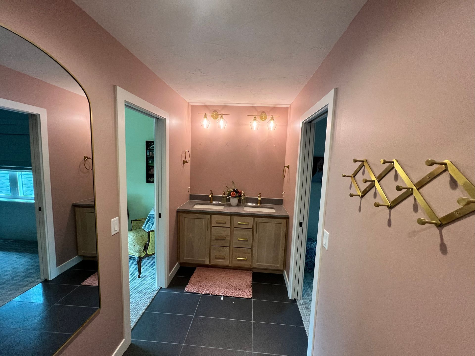 Pink-walled bathroom hallway with vanity, doors, gold coat rack, and large mirror. Dark tile floor.