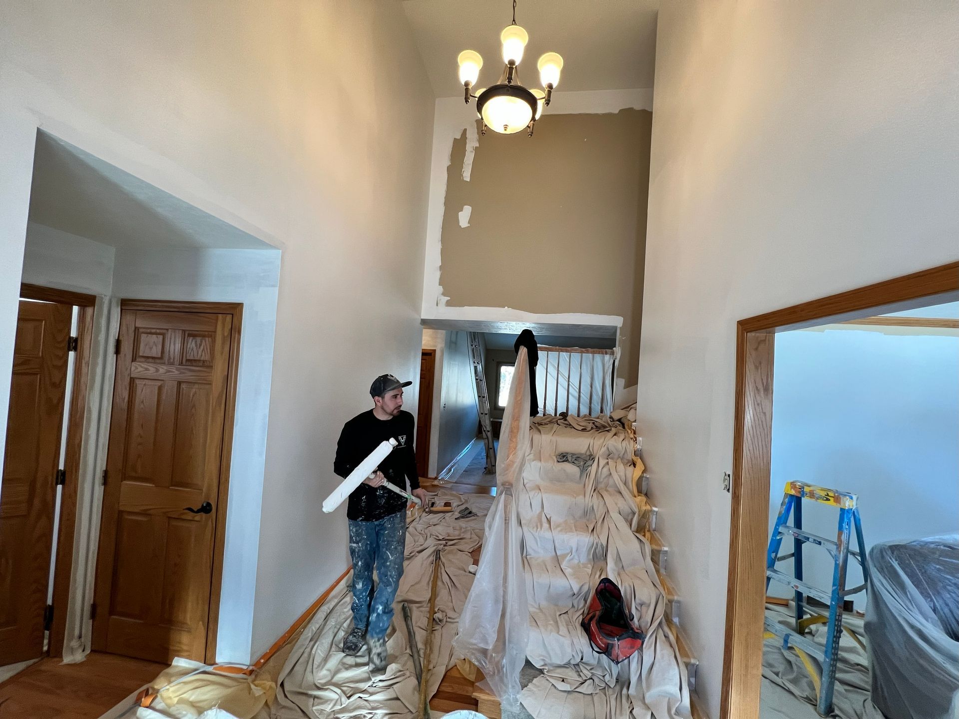 Person painting a stairwell in a home. The stairs are covered, and the walls are partially painted beige.