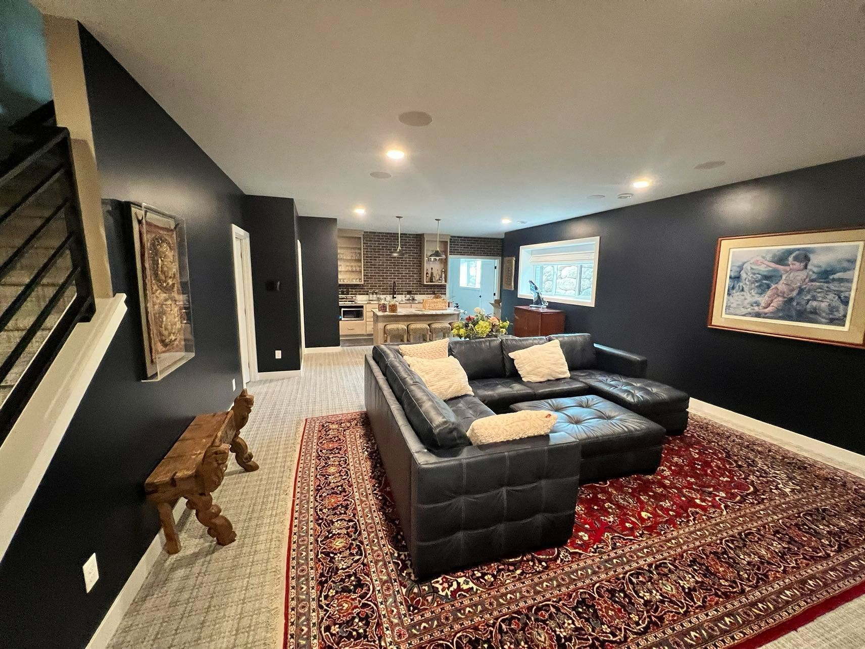 Dark-walled living room with sectional sofa on a Persian rug. Stairs, art, and a kitchen are visible in the background.