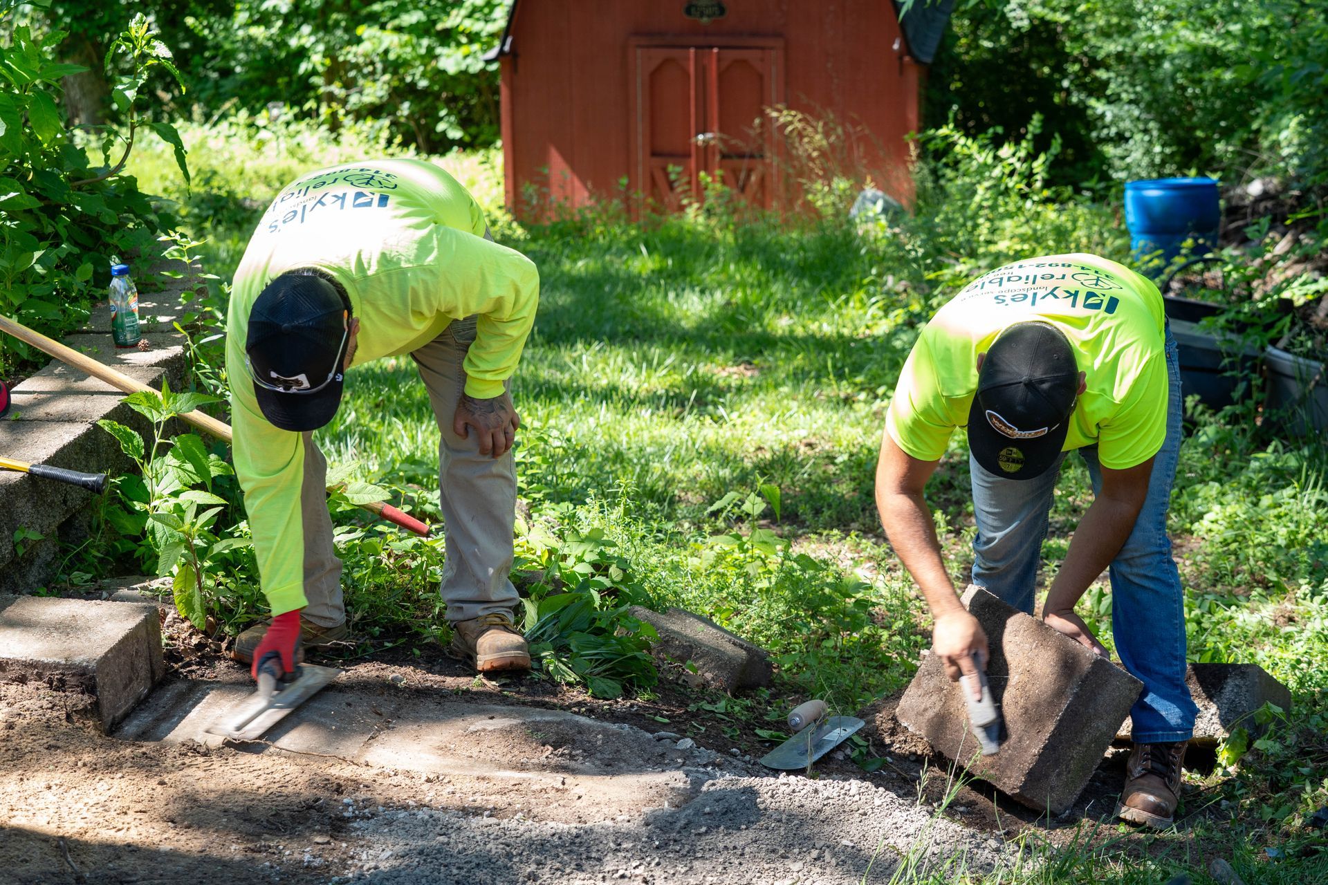 Two workers laying bricks in a grassy area, wearing neon yellow shirts and baseball caps.