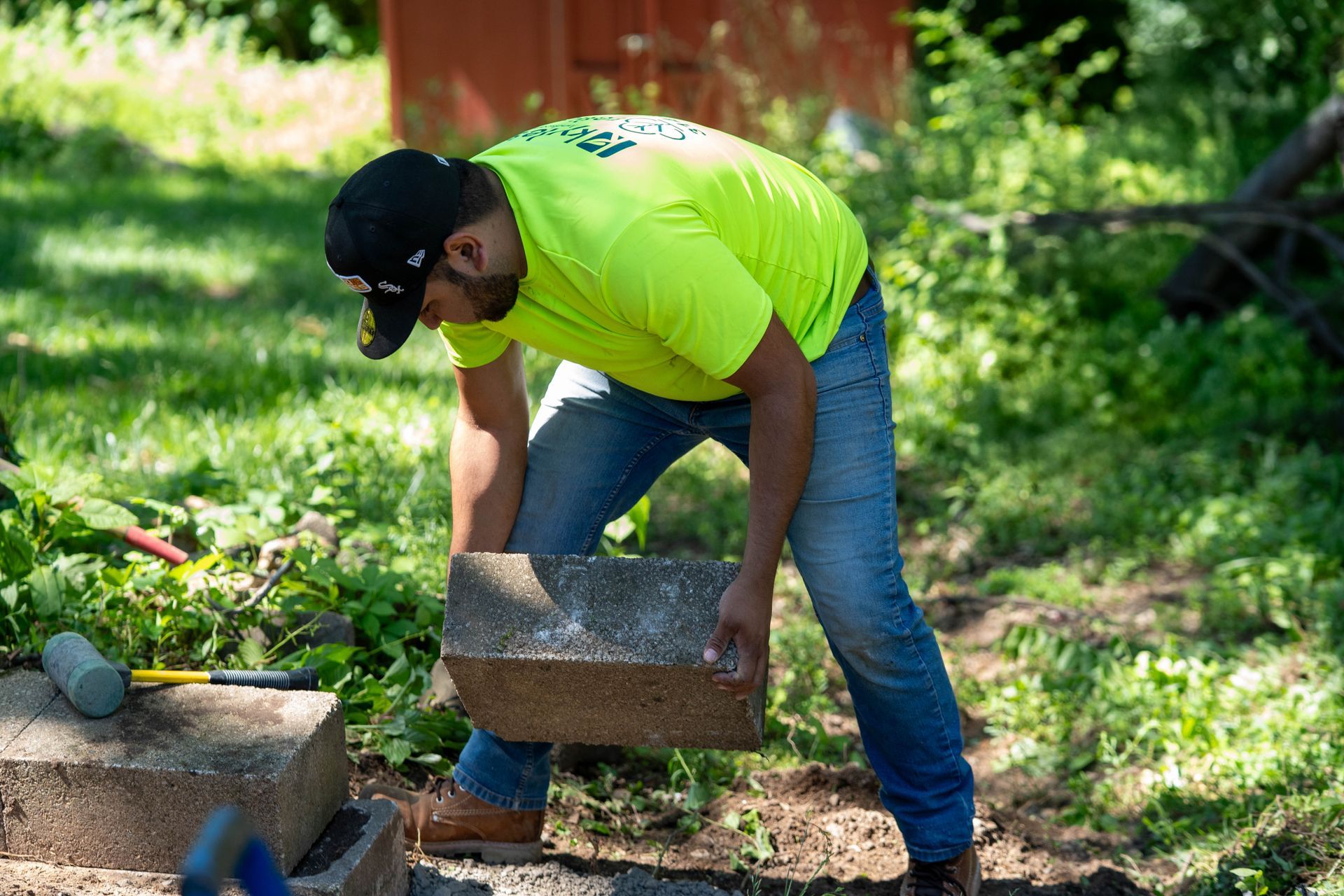 Man in neon green shirt and jeans lifting a stone block outdoors.