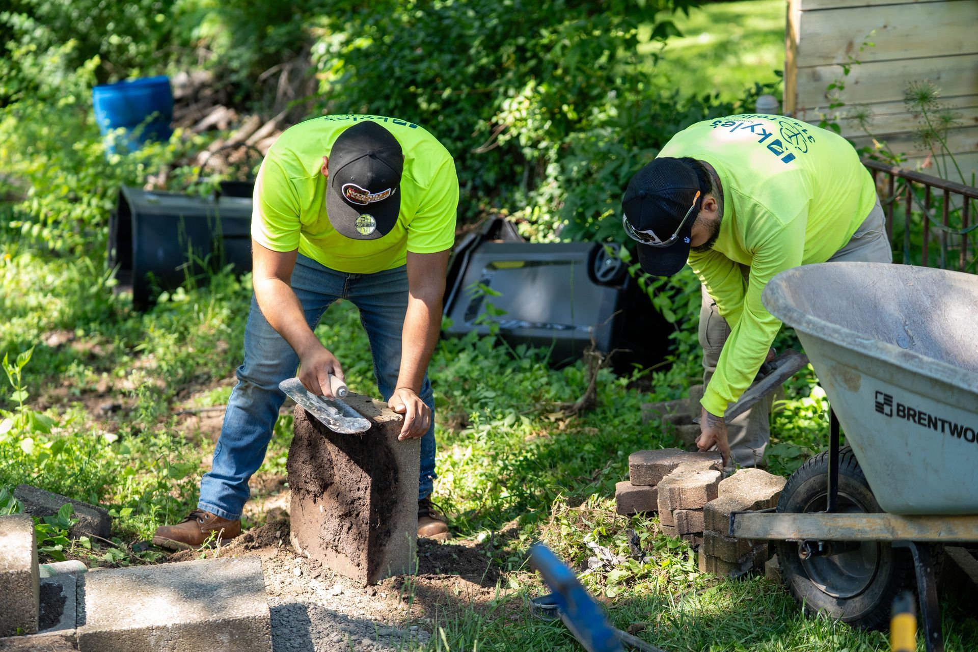 Two workers in neon green shirts laying bricks outdoors, with tools and a wheelbarrow.