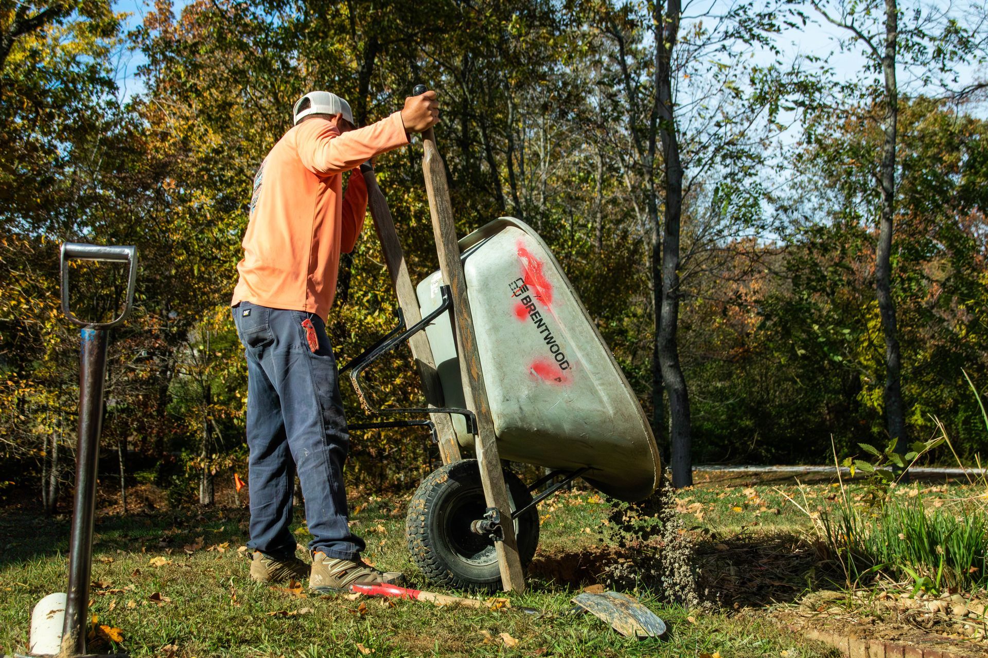 Person dumping contents from a wheelbarrow outdoors; trees in background.