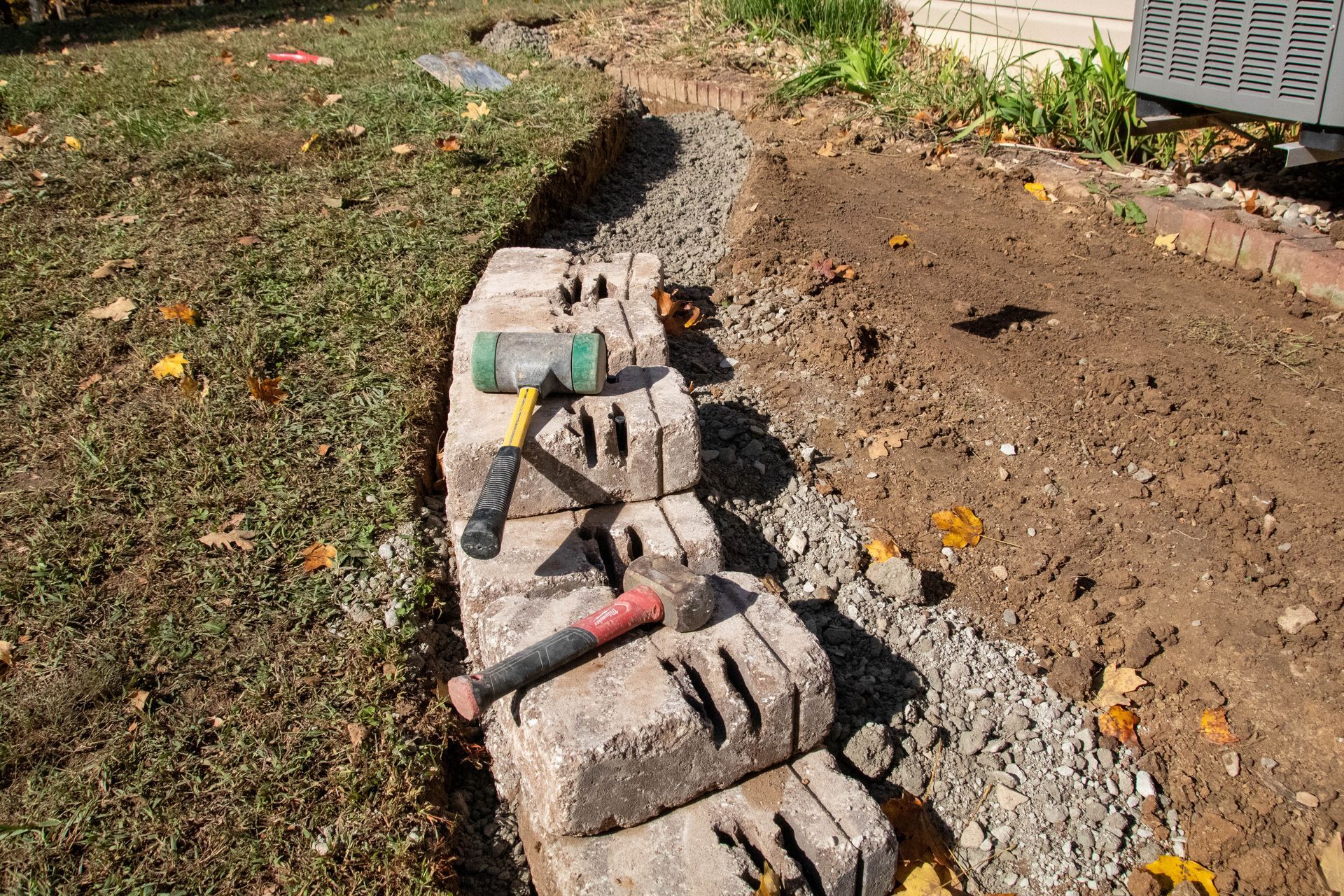 Building a retaining wall: bricks, gravel, and tools lie in a trench on a lawn.