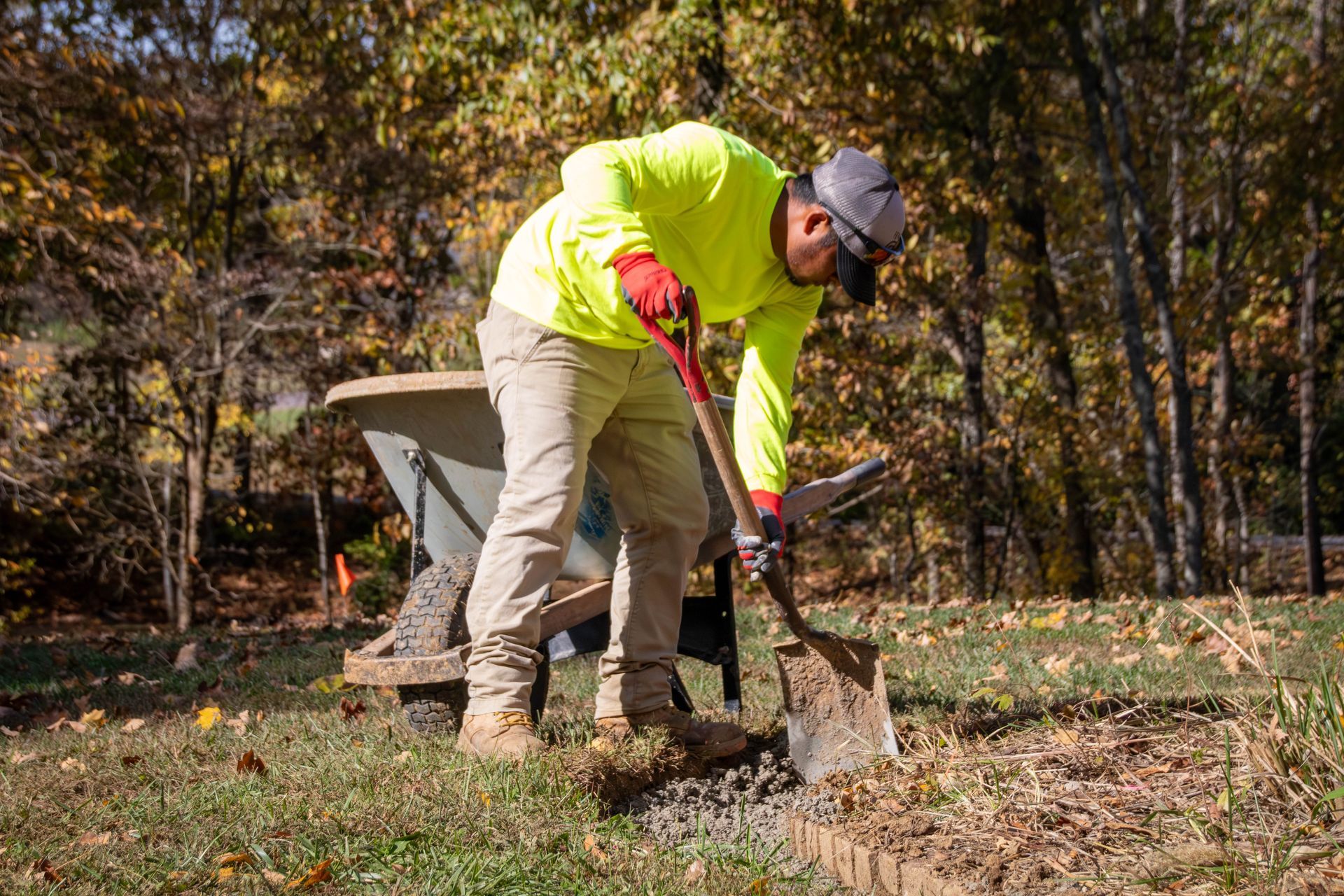 Person in neon shirt digging with a shovel next to a wheelbarrow, outdoors.