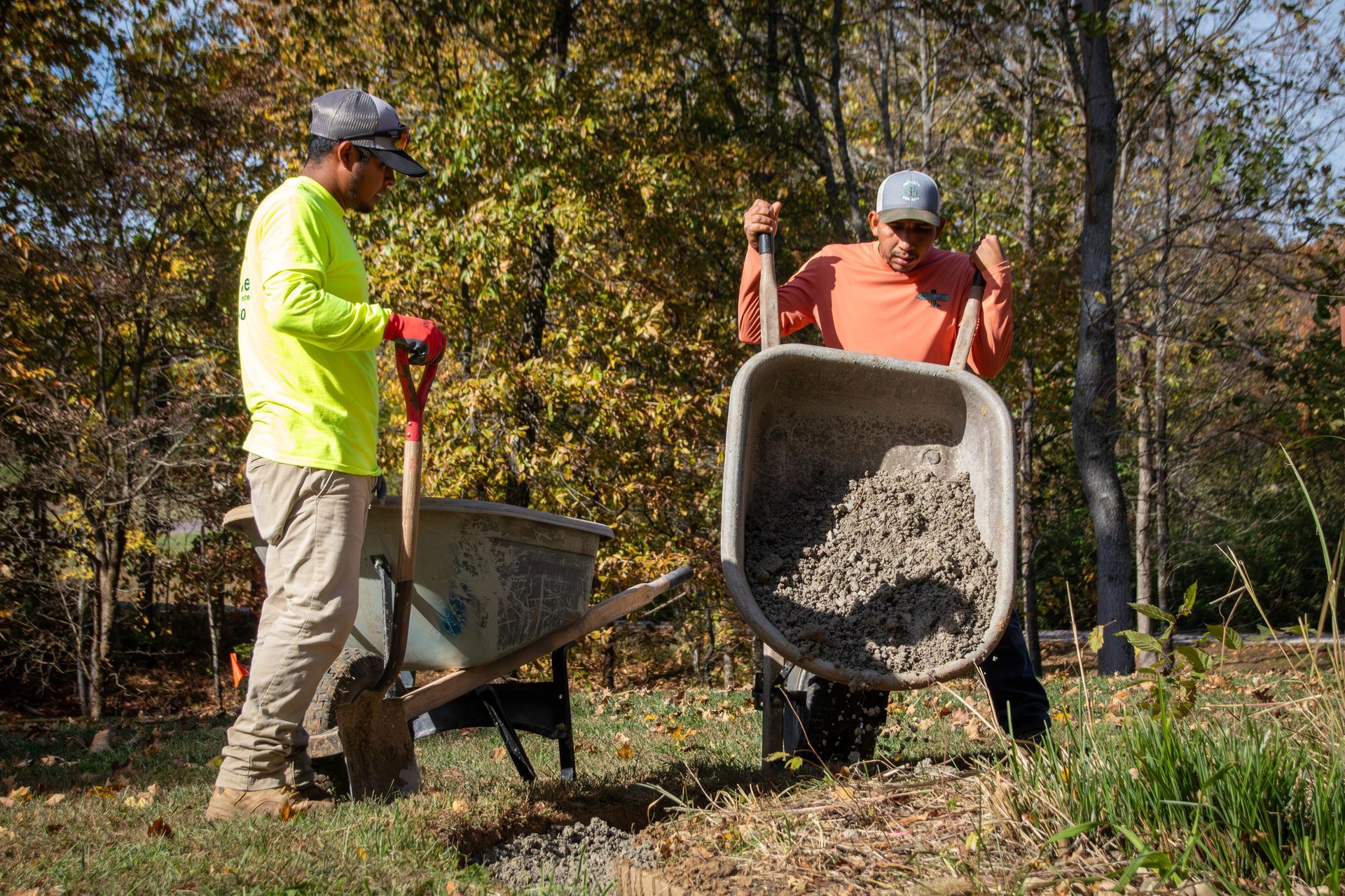 Two people filling a wheelbarrow with gravel in a grassy outdoor setting, one using a shovel.