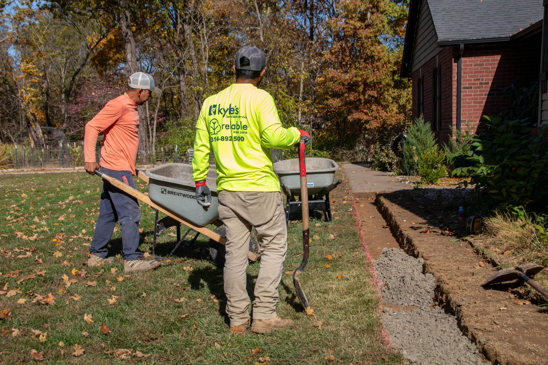 Two workers digging and moving dirt near a house; one uses a shovel, another a wheelbarrow. Sunny day.