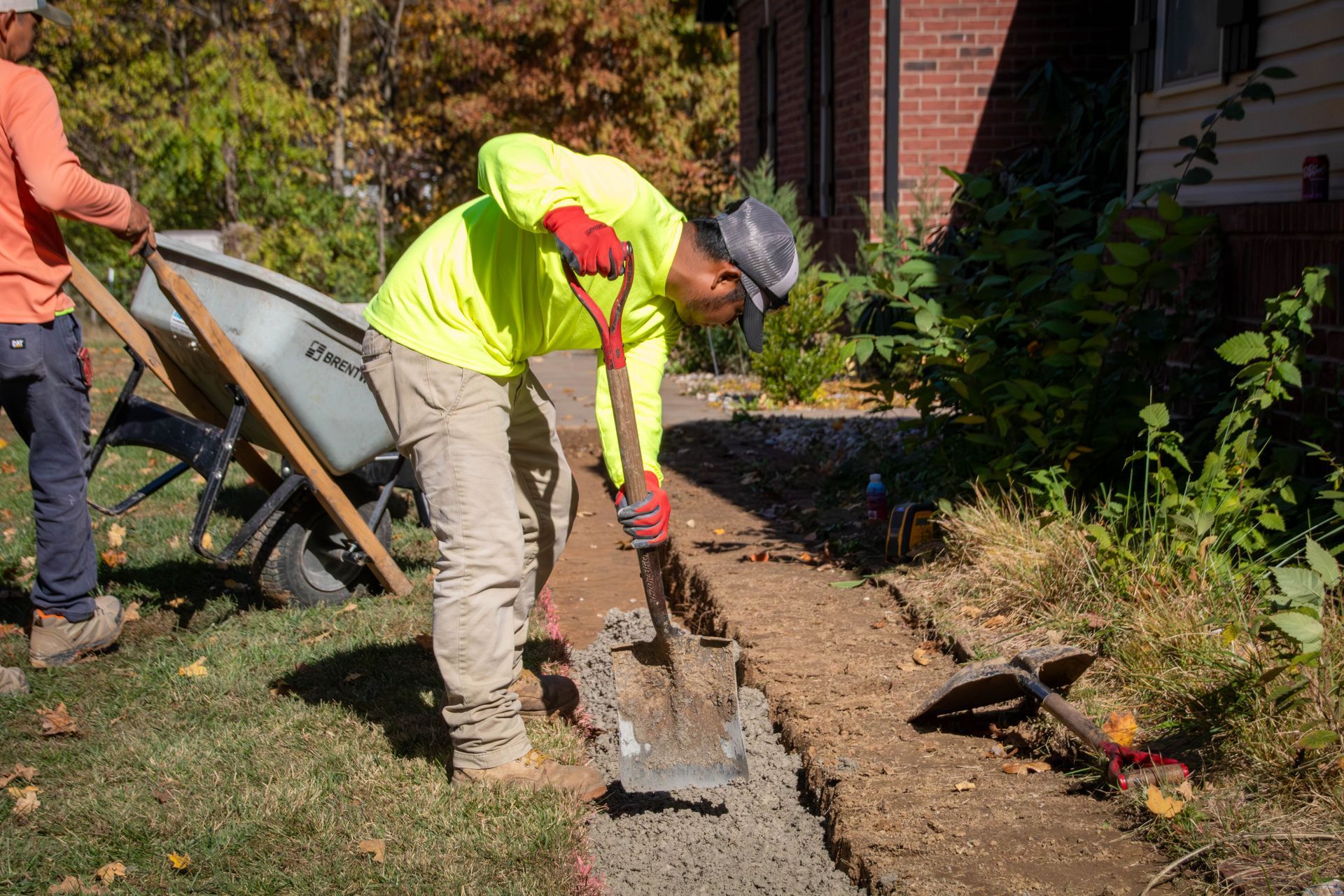Man in neon shirt using a shovel to pour concrete in a walkway, with a wheelbarrow and building in the background.