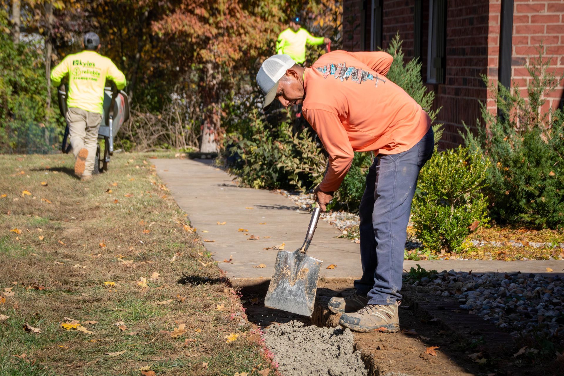 Three construction workers. One digs with a shovel, sidewalk and house visible, bright orange shirt.