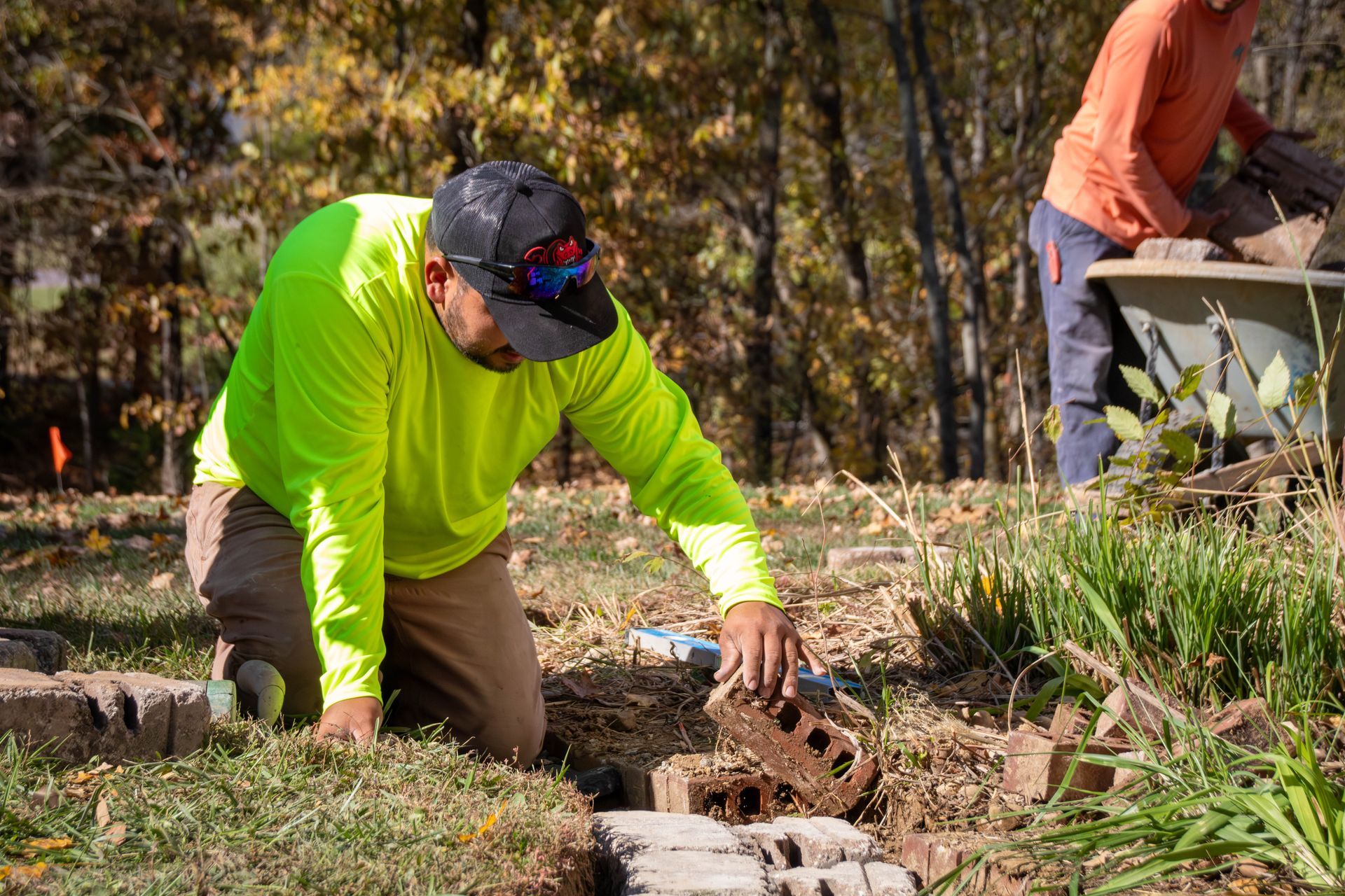 Two workers laying bricks outdoors; one kneels, placing a brick; bright sunlight.