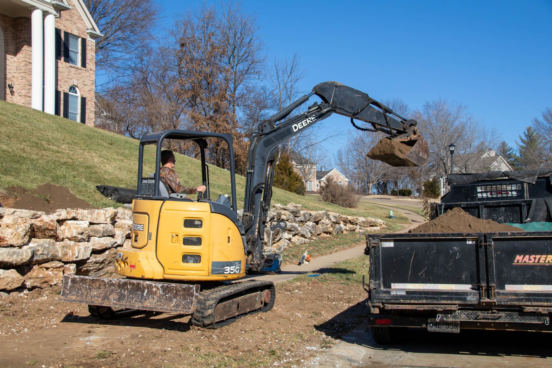 Yellow excavator loading dirt into a black dump truck next to a house with a rock retaining wall.