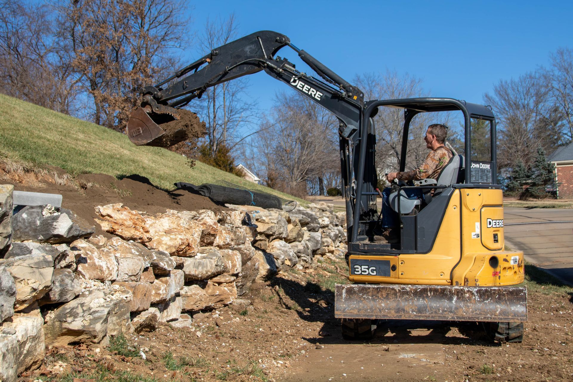 A small yellow excavator operated by a person is working on a rock wall. Sunny day.