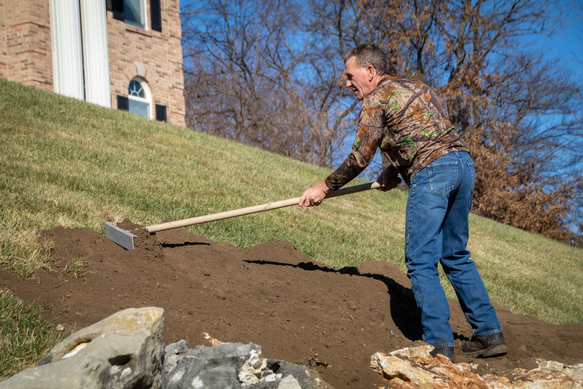 Man raking soil on a sloped yard near a house. He wears a camouflage shirt and jeans. Sunny day.