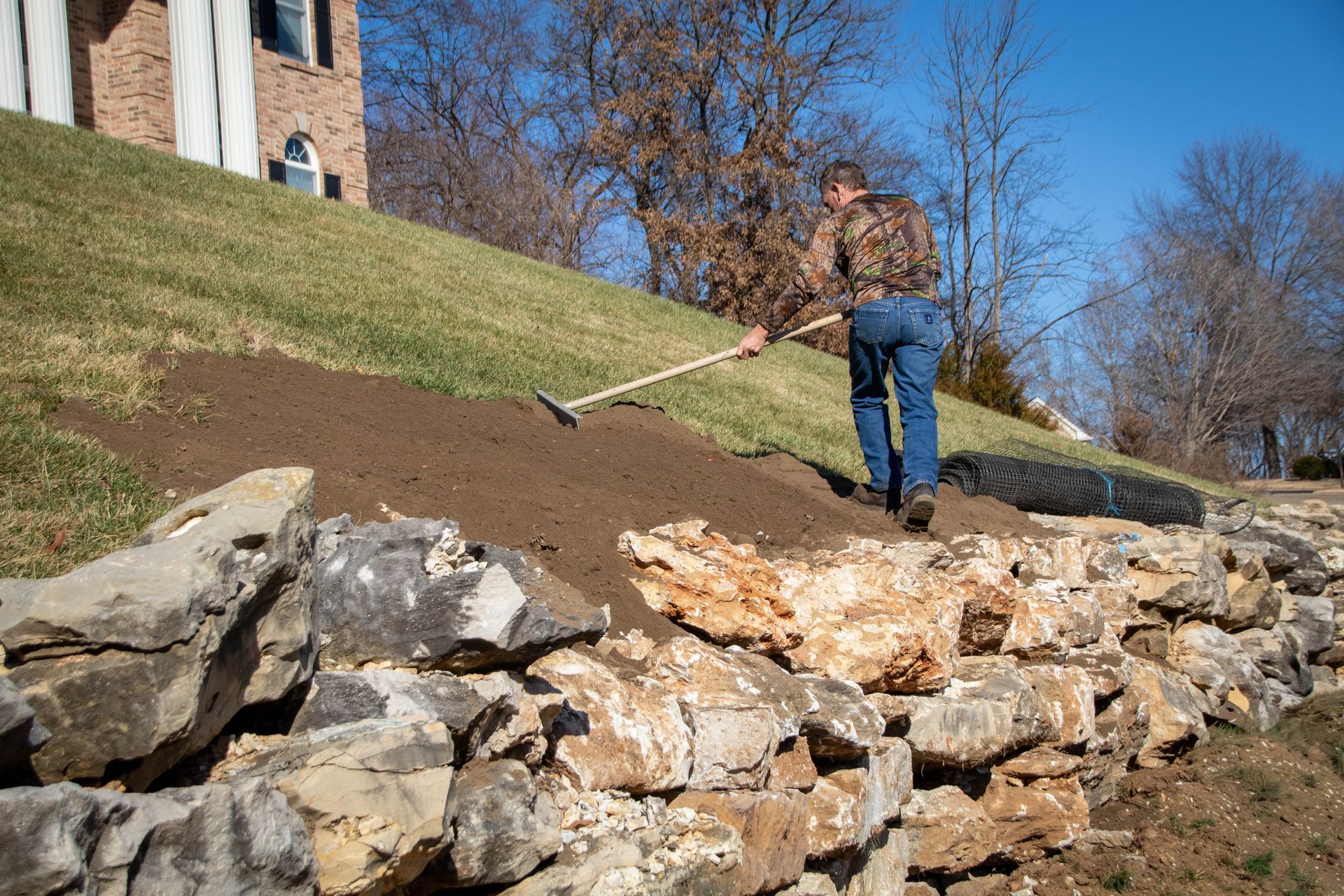 Man raking soil on a hillside, near a stone retaining wall and house.