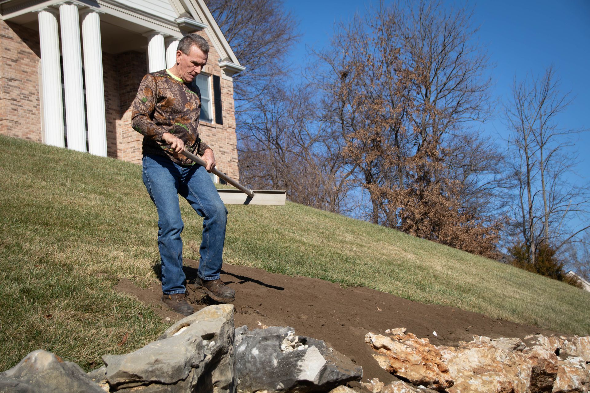 Man in camouflage shirt and jeans working on a hillside with a hoe. Near a house on a sunny day.